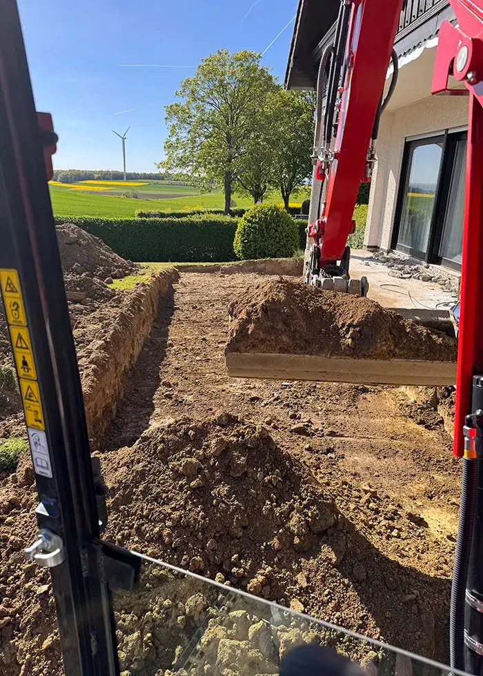 Baugrube neben einem Haus mit einem roten Bagger, der Erde aushebt, im Hintergrund Felder und ein Windrad