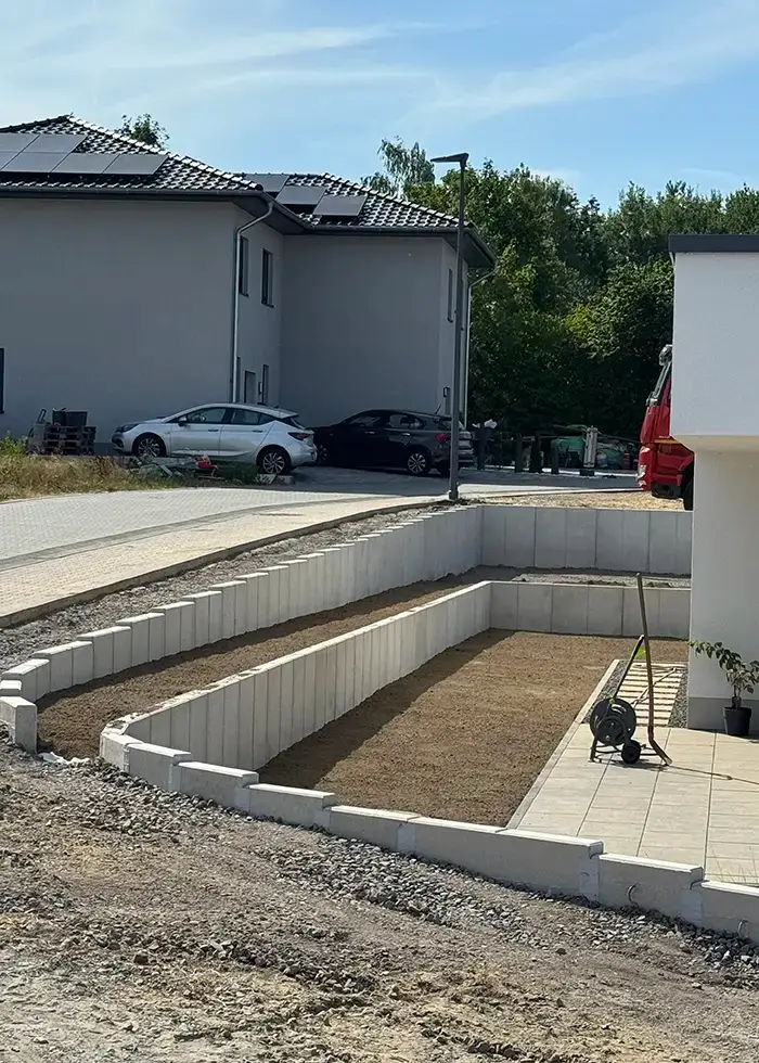 Terrasse mit gestuften Stützwänden aus Betonplatten neben einem weißen Haus und parkenden Autos im Hintergrund