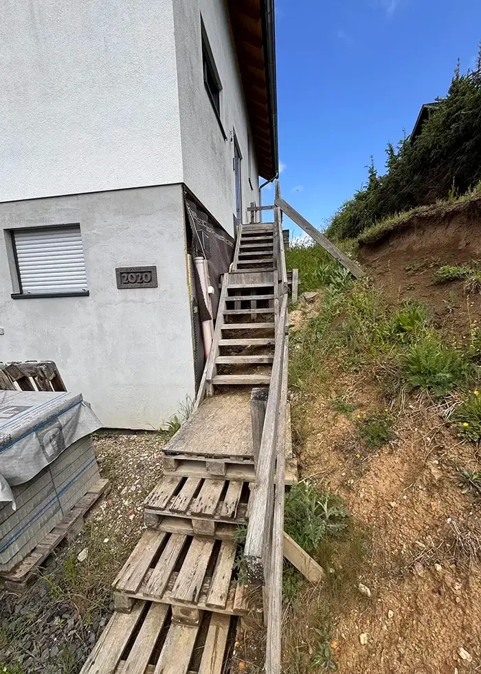 Holztreppe mit Geländer an einem Haus, rechts davon unbefestigter Hang mit Gras und Sträuchern