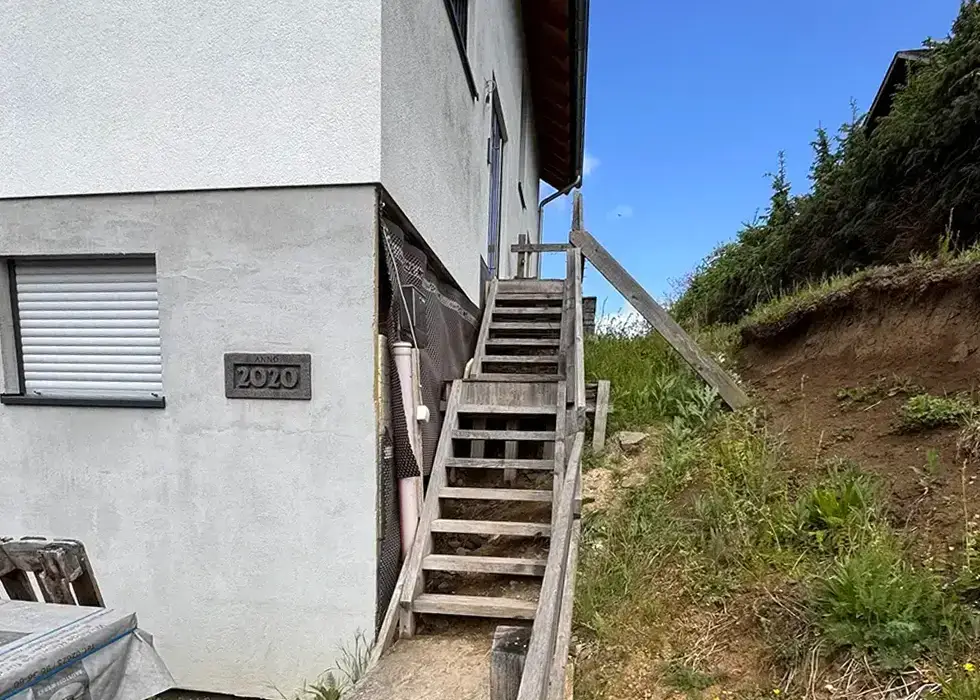 Holztreppe mit Geländer an einem Haus, rechts davon unbefestigter Hang mit Gras und Sträuchern