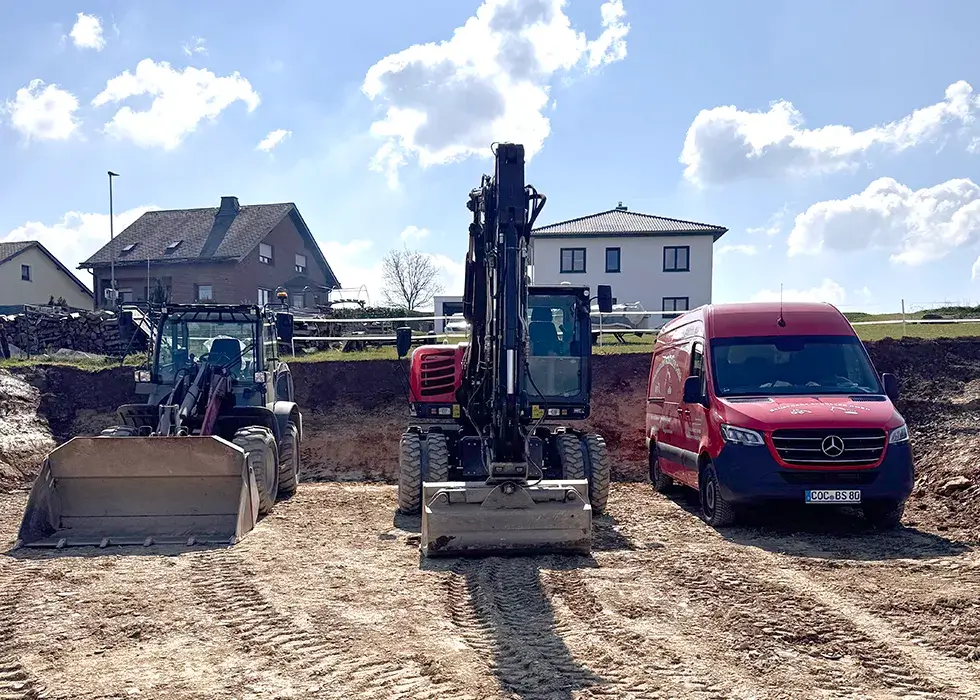 Bauplatz mit einem Radlader, einem Bagger und einem roten Mercedes-Transporter nebeneinander auf unbefestigtem Boden vor Wohnhäusern