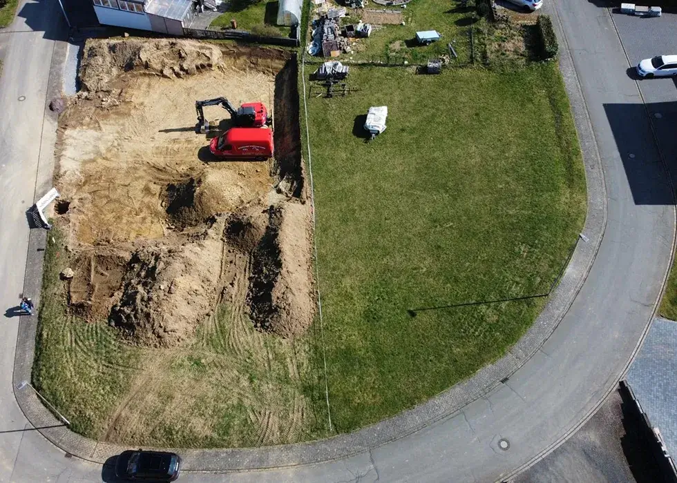 Luftaufnahme einer Baustelle mit einem roten Bagger neben einer grünen Wiese und angrenzenden Straßen und Gebäuden