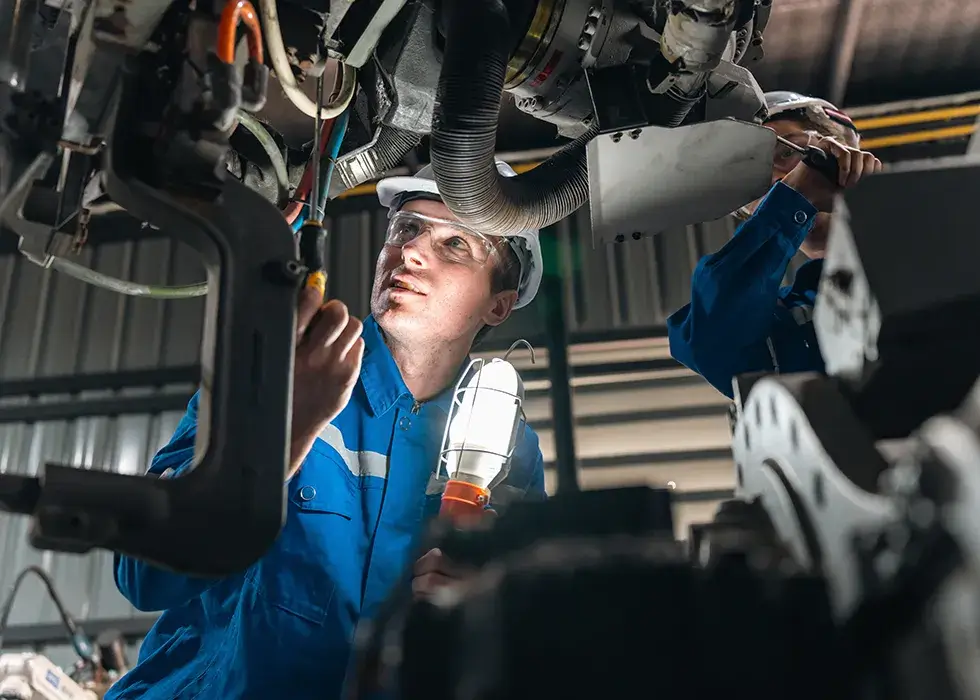 Techniker mit Schutzbrille und blauem Overall inspiziert eine Maschine in einer Werkstatt und hält eine Arbeitslampe