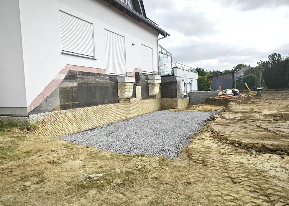 Haus mit abgetragenem Erdboden und Schotterfläche vor der Fassade unter geschlossenen Rollläden